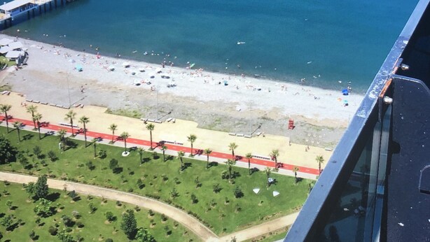 An aerial view of a beach with a pier extending into the water, a sandy beach with umbrellas, and a road with palm trees. - Hotel 36 - ORBI Beach Tower Batumi 36th floor (Batumi)