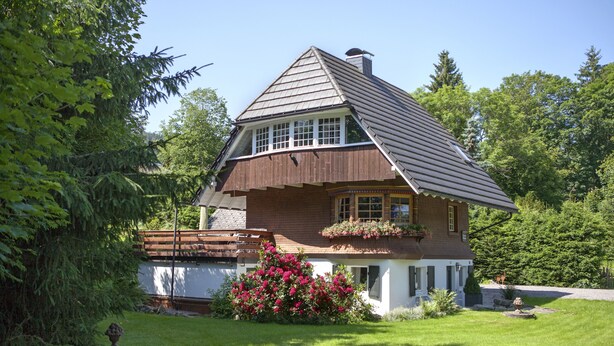 A two-story house with a sloped roof, a wooden deck, and a garden with flowering plants. - Chalet am Birkenhain (Hinterzarten)