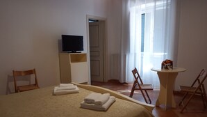 A hotel room with a bed, a TV on a stand, a small table with a vase, and two chairs. - Lamione da Dorotea (Torchiara)