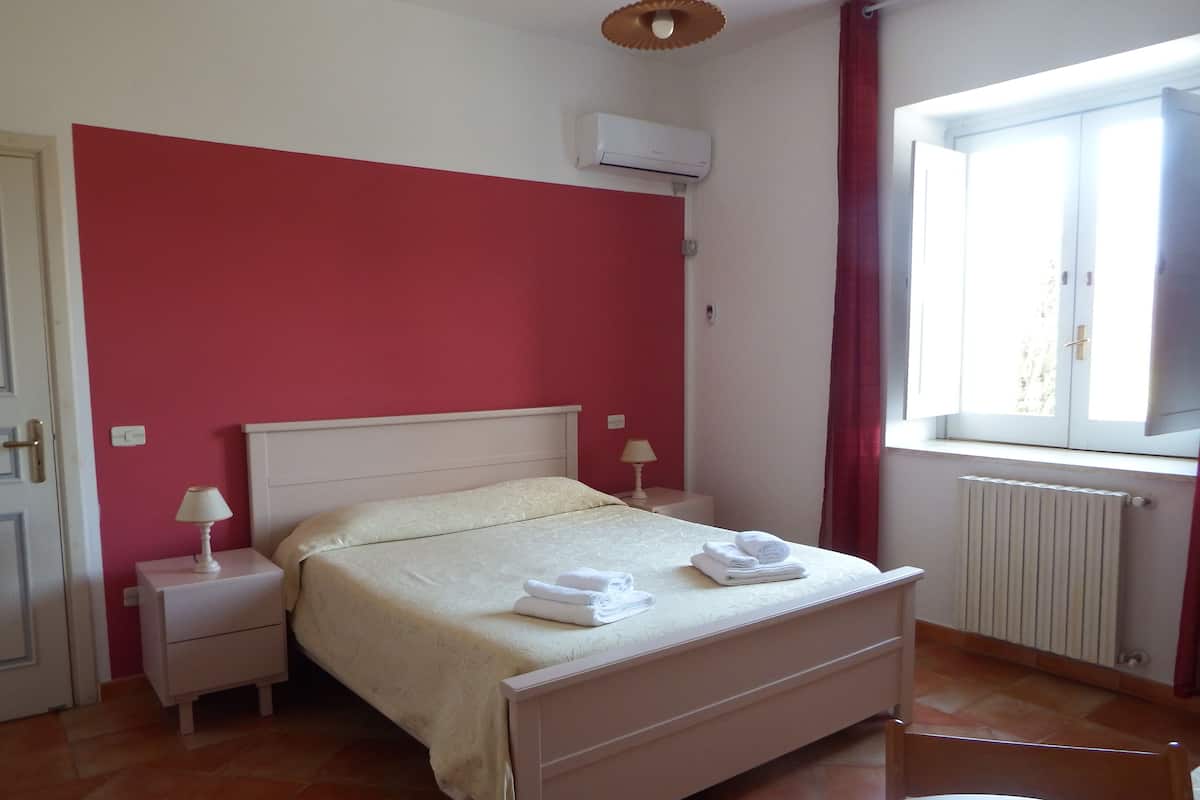 A bedroom with a bed, two bedside tables with lamps, a window with curtains, and a radiator.