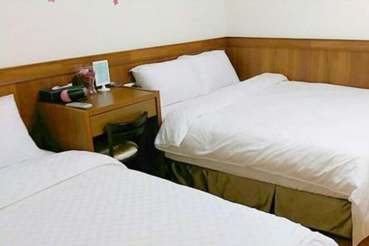 A hotel room with two single beds, a wooden headboard, a bedside table with a lamp, and a small desk area.