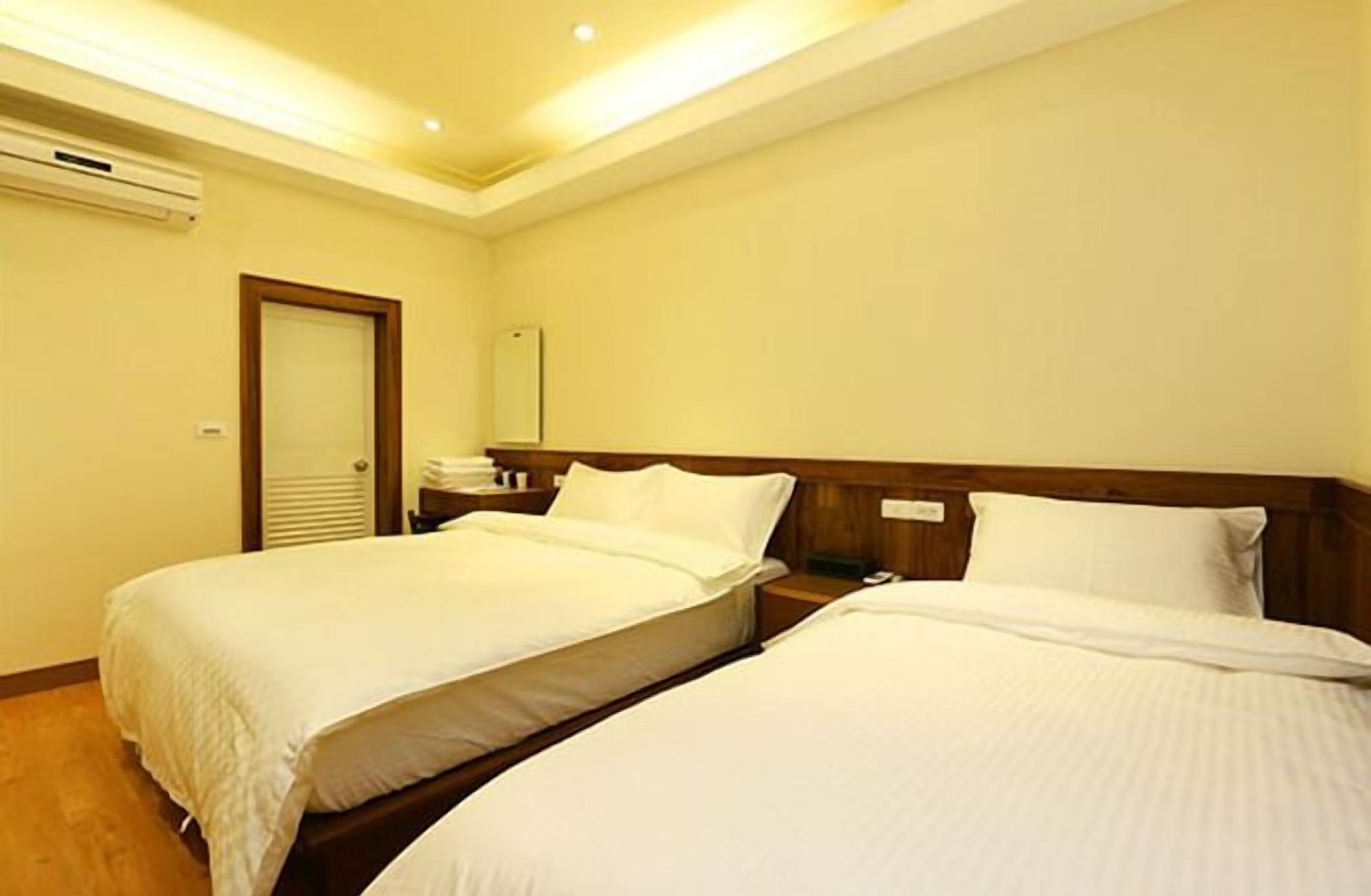 A hotel room with two single beds, a wooden headboard, a bedside table, and a wall-mounted air conditioning unit.