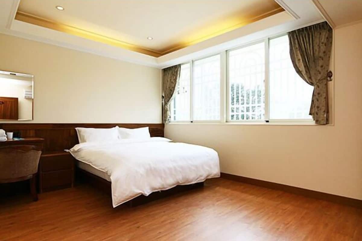 A hotel room with a wooden floor, a large bed, a bedside table, a chair, and a window with curtains.