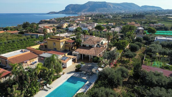 An aerial view of a coastal residential area with houses, a swimming pool, and a view of the sea and mountains in the background.