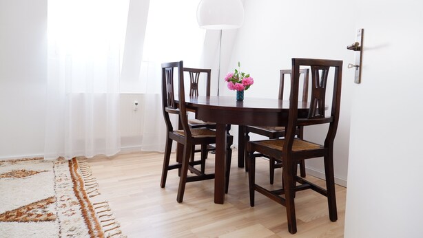 En matplats med ett runt träbord, fyra mörka trästolar, en vas med rosa blommor, en vit golvlampa och en vit vägg.