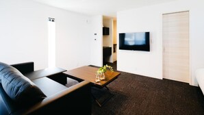 A modern living room with a black leather sofa, a wooden coffee table with a vase of flowers, a flat-screen TV mounted on the wall, and a sliding door leading to another room. - FUKUOKA MOJIKO Stay (Kitakyushu)