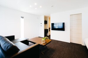 A modern living room with a black leather sofa, a wooden coffee table with a vase of flowers, a flat-screen TV mounted on the wall, and a sliding door leading to another room. - FUKUOKA MOJIKO Stay (Kitakyushu)