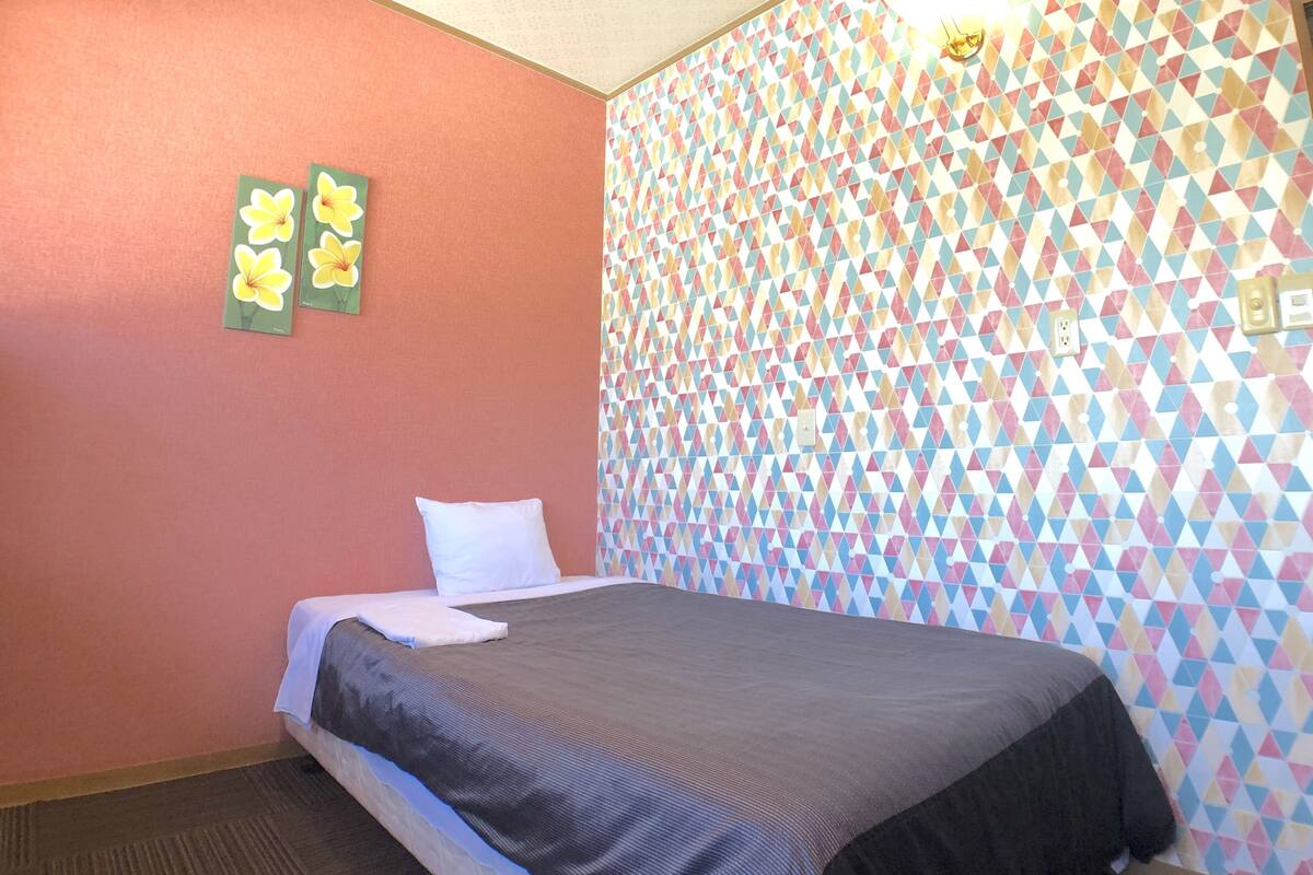 Una habitación de hotel con una cama, un almohadón, una pared con diseño geométrico y dos cuadros de flores.