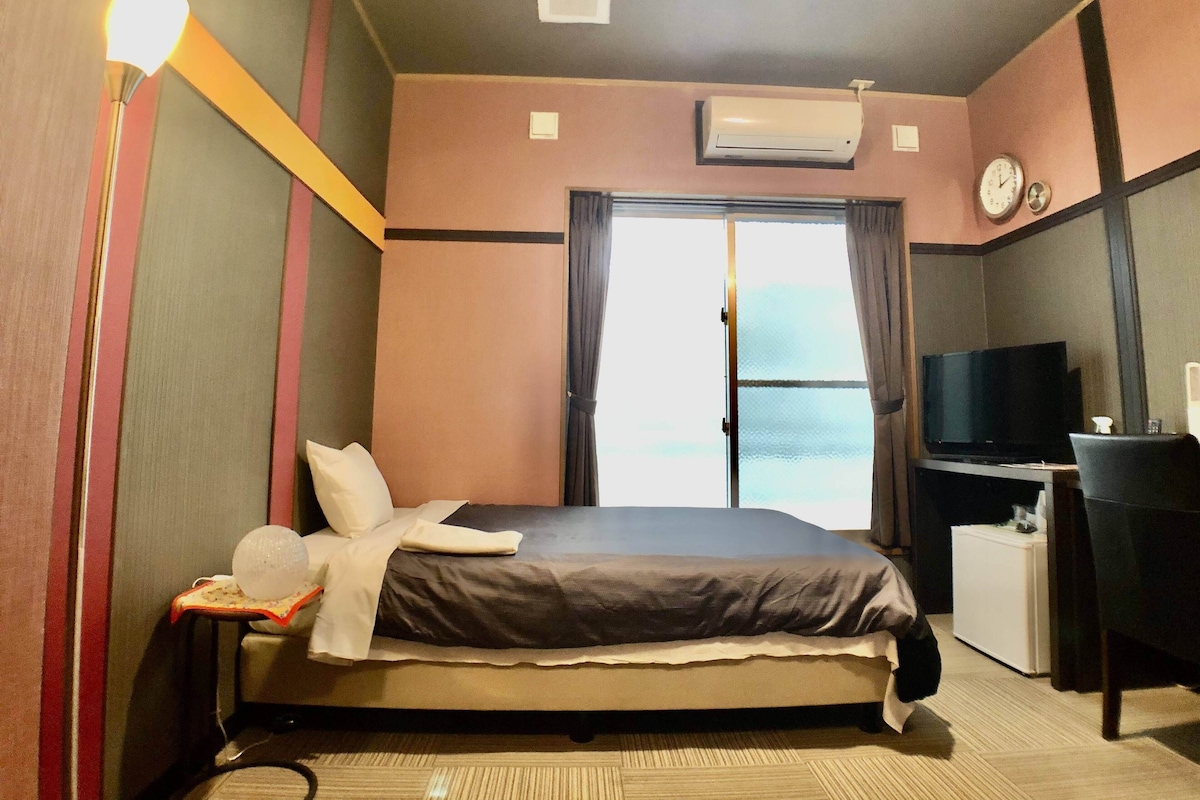 A hotel room with a bed, a desk with a television, an air conditioning unit, and a window with curtains.