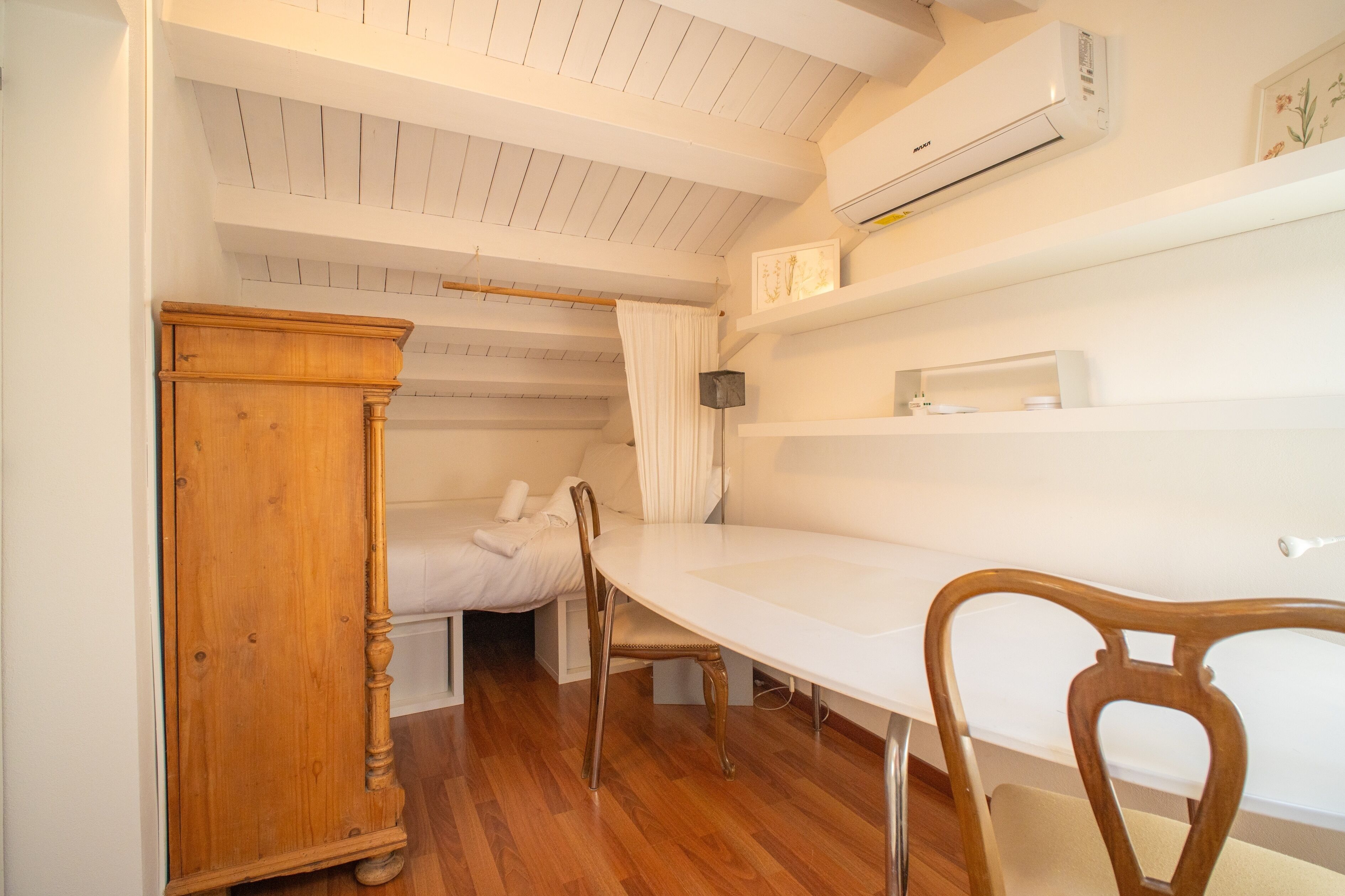 A compact room with a bed, a wooden dresser, a white table, and a chair.