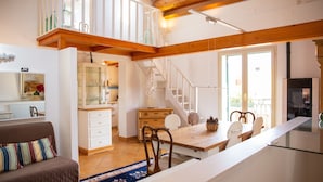 A spacious living area with a high ceiling, wooden beams, a dining table with chairs, a sofa with a patterned cushion, and a kitchen area with cabinets and a staircase. - Solemar Sicilia - Casa Olivia (Santa Flavia)