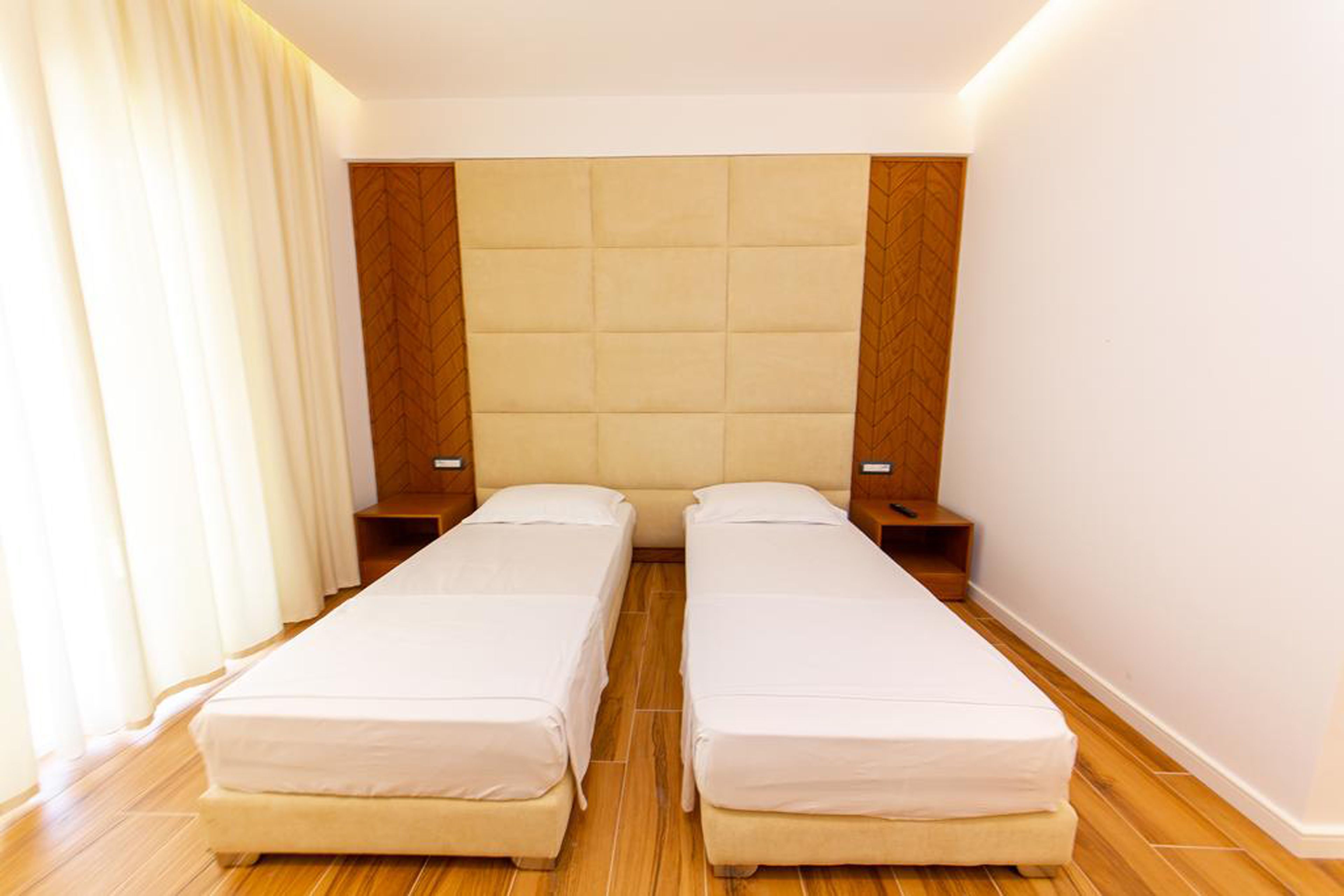 Habitación doble con dos camas individuales, cabecero de madera, mesitas de noche y un ventanal con cortinas.