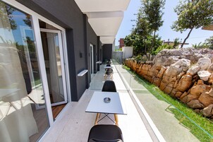Un área de estar moderna al aire libre con mesas y sillas, un acceso por puerta de vidrio y una pared de piedra.