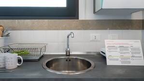 A stainless steel kitchen sink with a faucet, a metal dish rack with utensils, and two white mugs with the 'Italianway' logo. A recycling information sign is visible on the wall. - Italianway - Sant'Oronzo Apartment (Lecce)