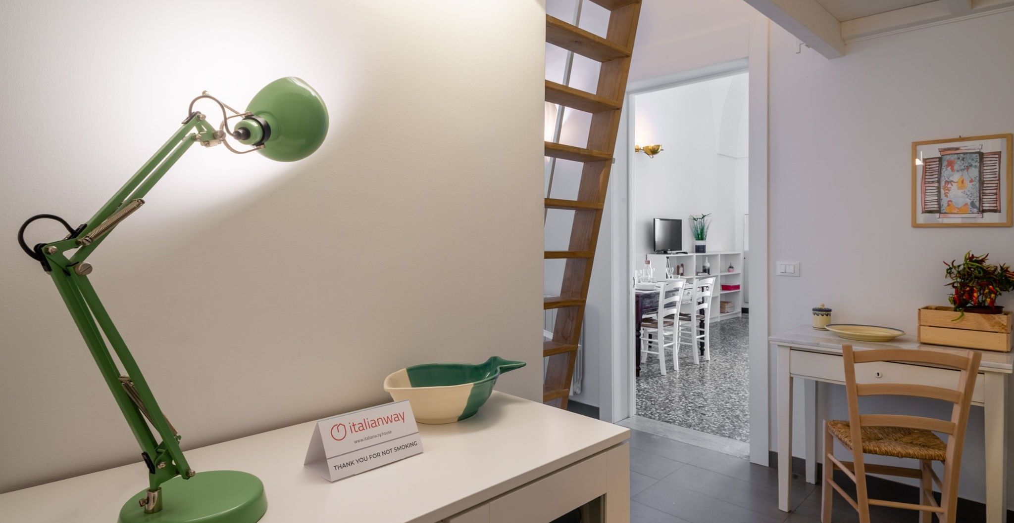 A room with a desk, a green desk lamp, a sign, a chair, and a door leading to a dining area.