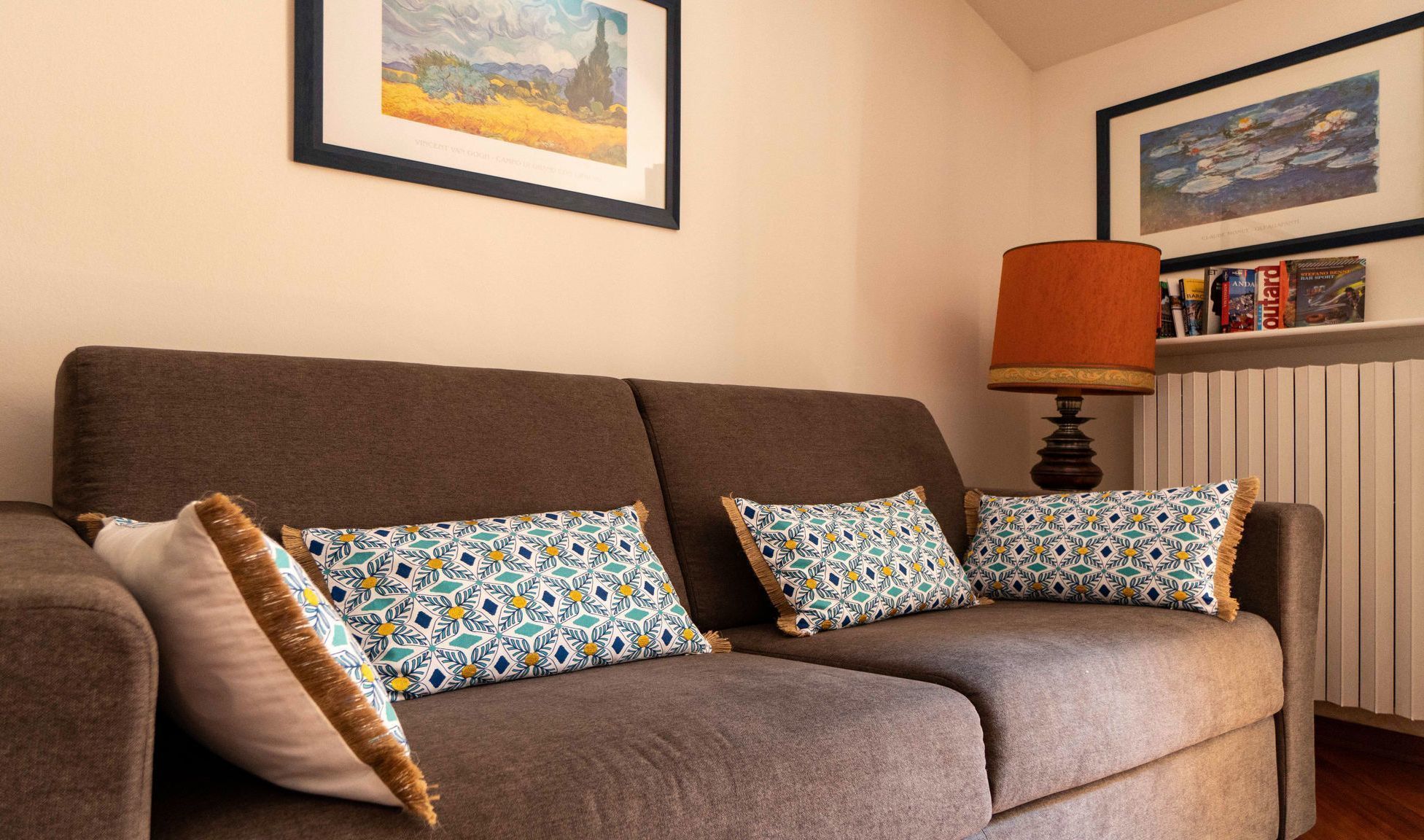 A brown sofa with patterned cushions, a framed painting above, and a lamp on a side table.