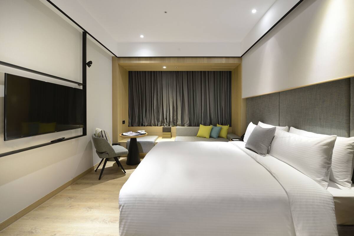 Una habitación de hotel moderna con una cama grande, un televisor de pantalla plana montado en la pared, una pequeña mesa redonda con una silla y una ventana con cortinas.
