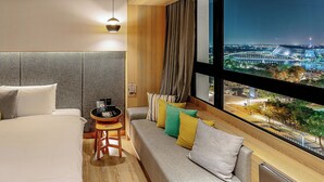 Habitación de hotel moderna con una cama grande, un sofá, una mesita con dispositivos electrónicos y vistas a un paisaje urbano nocturno.