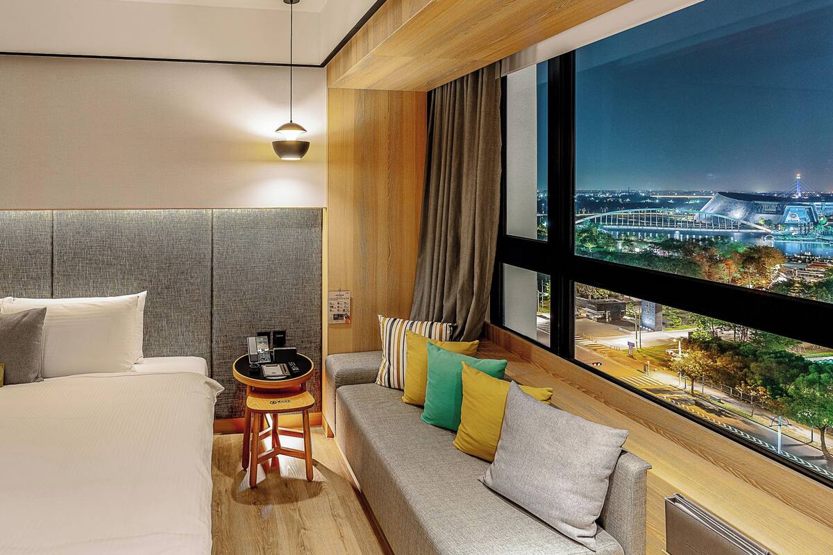 Habitación de hotel moderna con una cama grande, un sofá, una mesita con dispositivos electrónicos y vistas a un paisaje urbano nocturno.