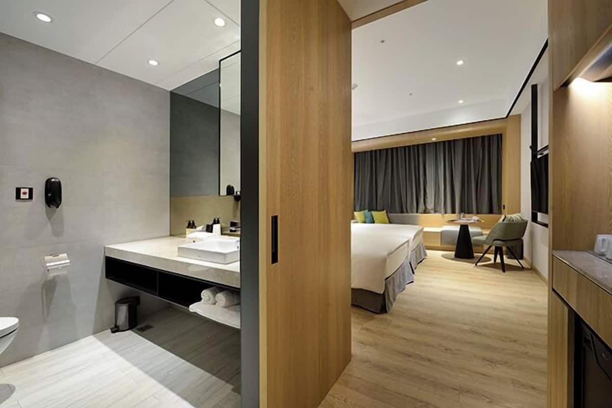 Una habitación de hotel moderna con piso de madera, una cama grande, una mesita de noche, una silla, un escritorio y un baño con lavamanos y espejo.
