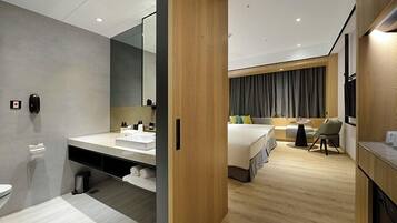 A modern hotel room with a wooden floor, a large bed, a bedside table, a chair, a desk, and a bathroom with a sink and mirror.