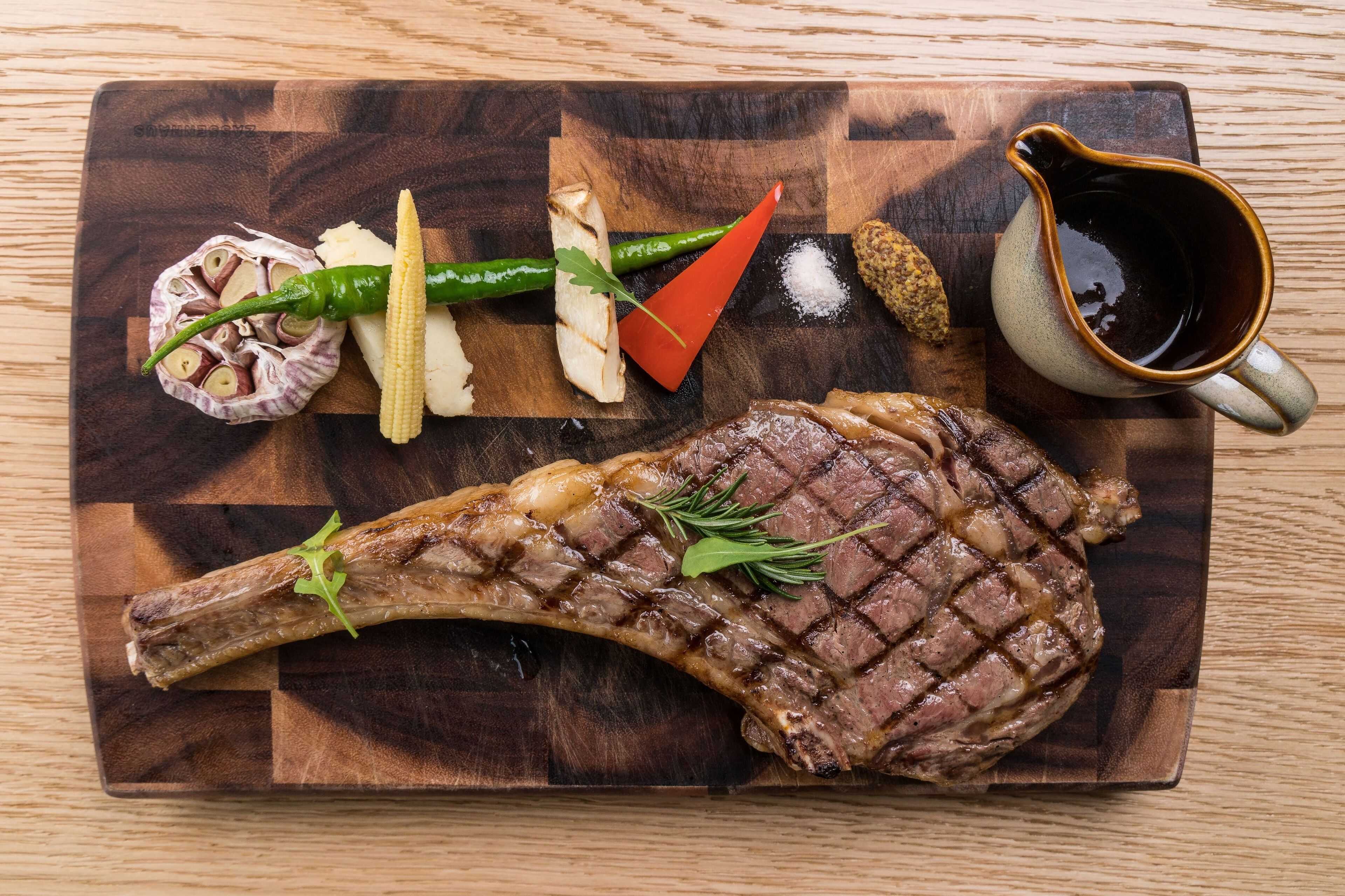 A steak with vegetables and a sauce pitcher on a wooden board.