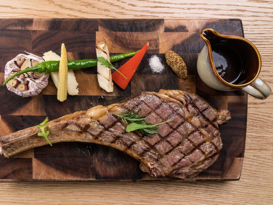 A steak with vegetables and a sauce pitcher on a wooden board.