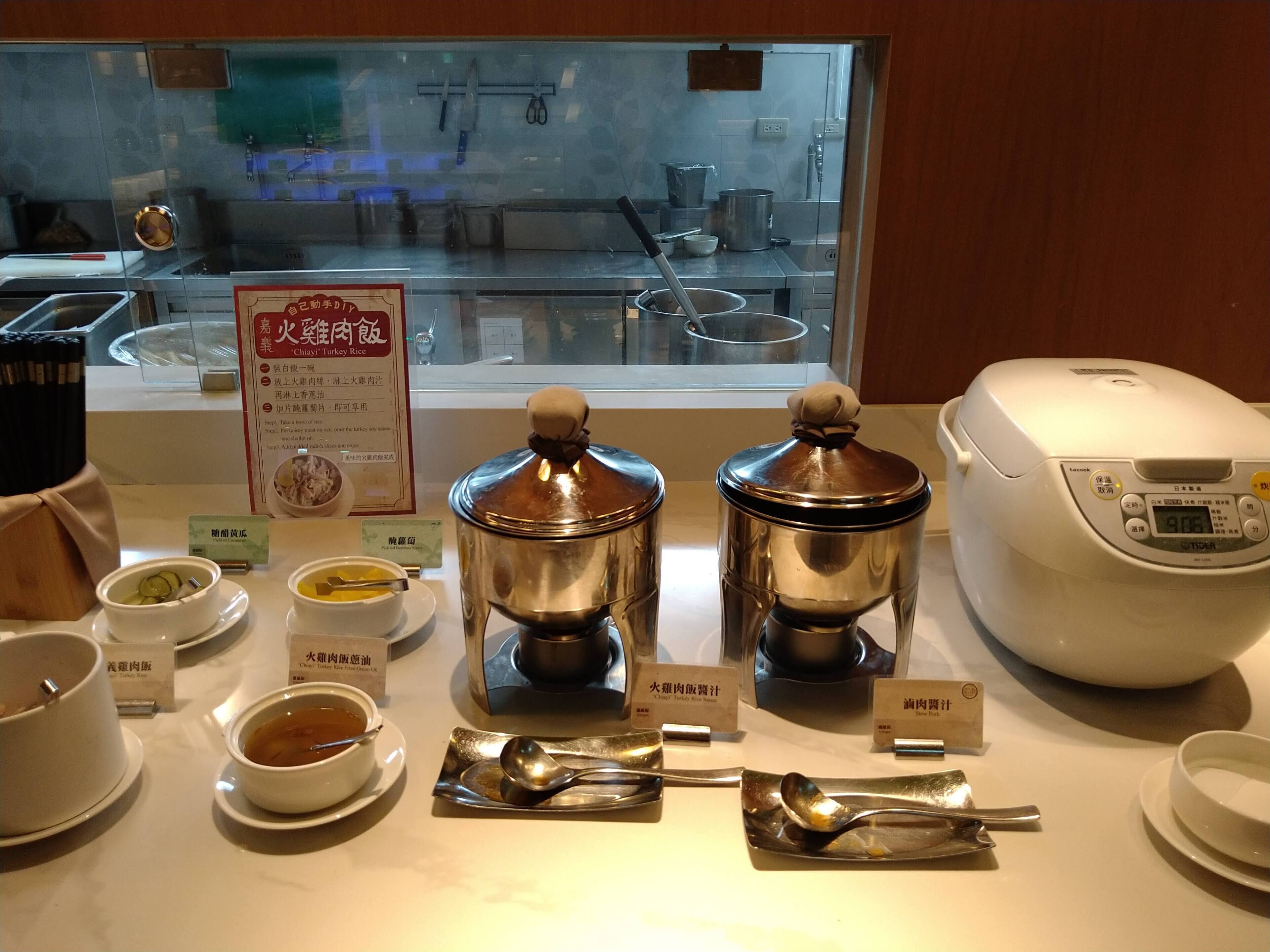Una zona de cocina con recipientes de acero inoxidable etiquetados para distintos platos, una cocedora de arroz y utensilios sobre una encimera.