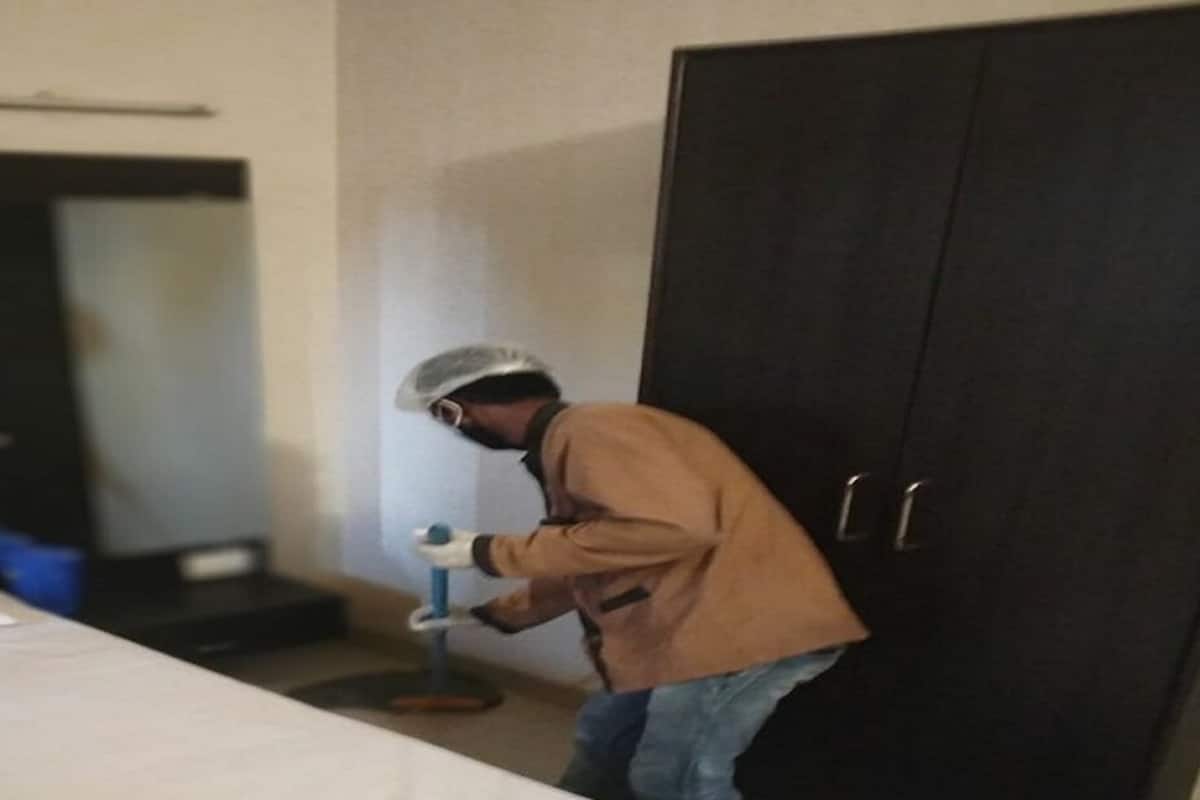A person in a brown jacket and jeans, wearing a hairnet, standing in a room with a bed, a dark wardrobe, and a small table with a blue object on it.