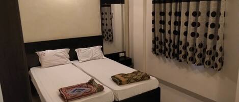 A double bed with white linens, a black headboard, and two pillows. There is a small bedside table with a lamp. The room has a mirror above the bed and a curtain with a polka dot pattern.