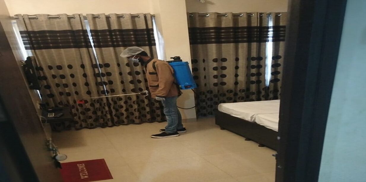 Een kamer met een bed, gordijnen met stippen, een rode welkomstmat en een persoon met een rugzak en een schoonmaakmiddel.