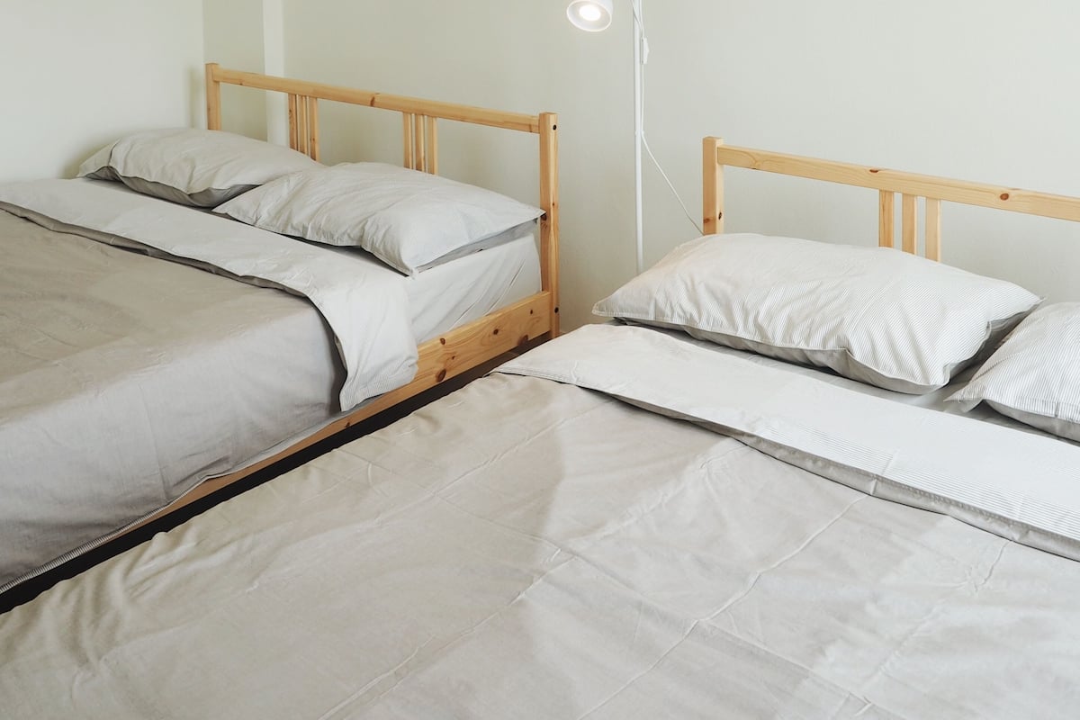 A room with two wooden beds, each with a white mattress and pillows. There is a white floor lamp on the right side of the image.
