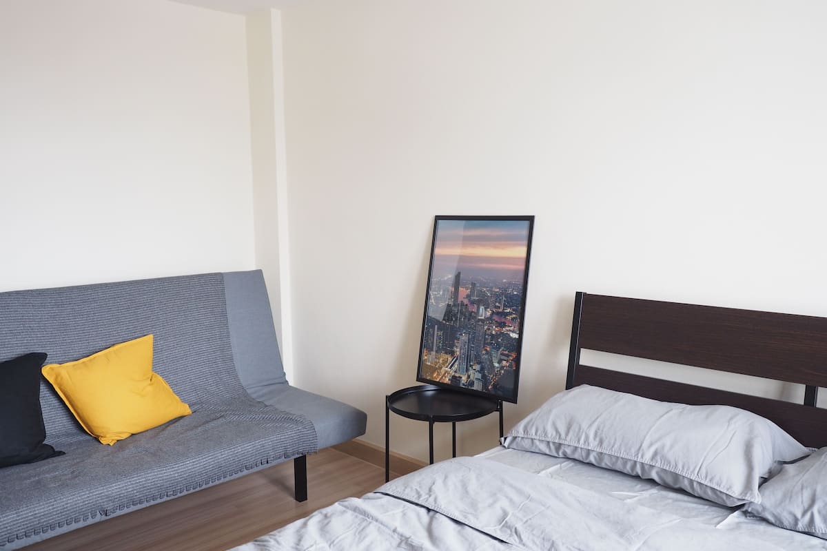 A hotel room with a grey sofa, a small round table, a bed with grey bedding, and a cityscape painting on the wall.