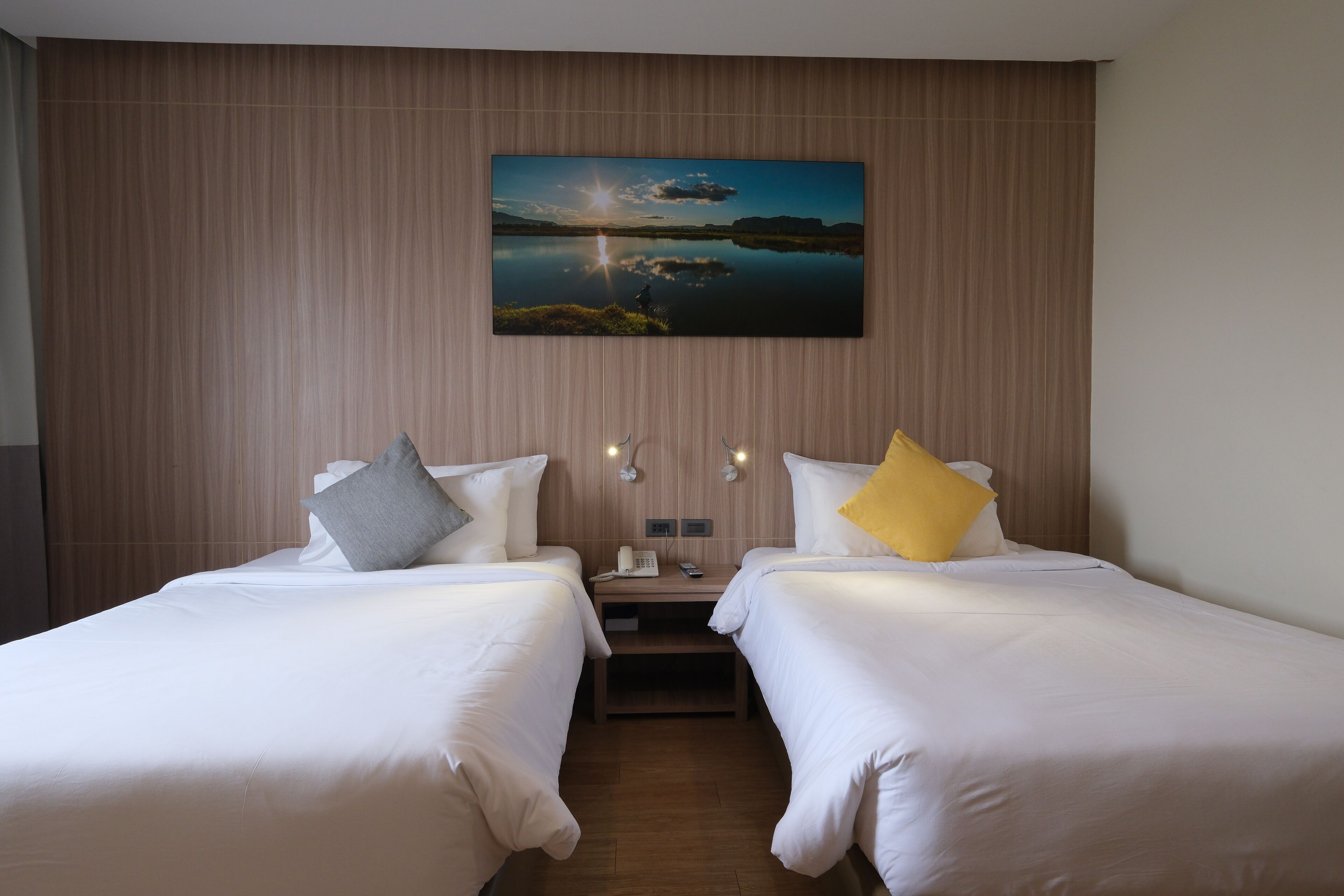 Habitación de hotel con dos camas, una mesita de noche, una lámpara y un cuadro de un paisaje.
