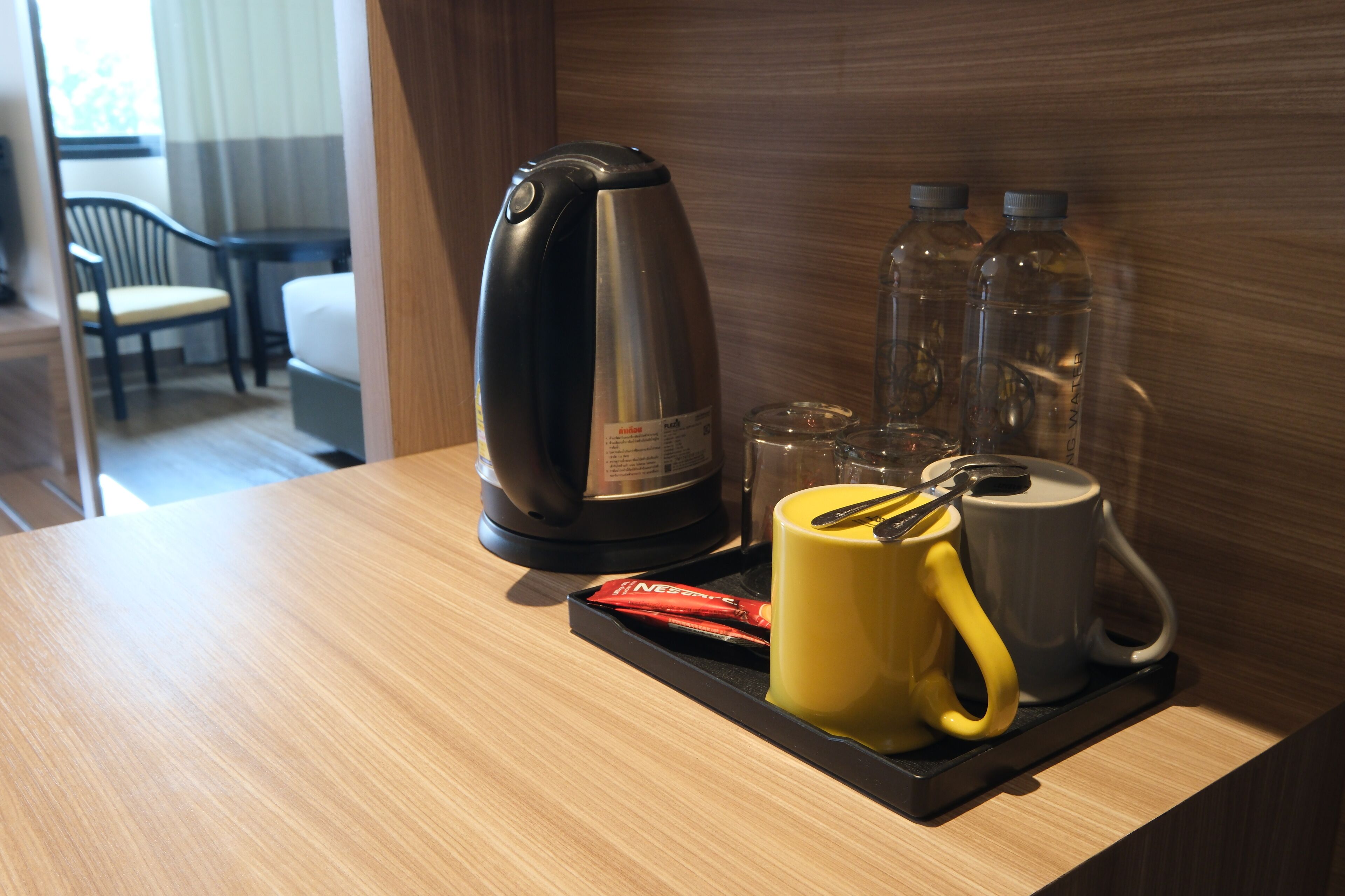 A hotel room with a wooden table, a coffee maker, a yellow mug, a white mug, and two water bottles.