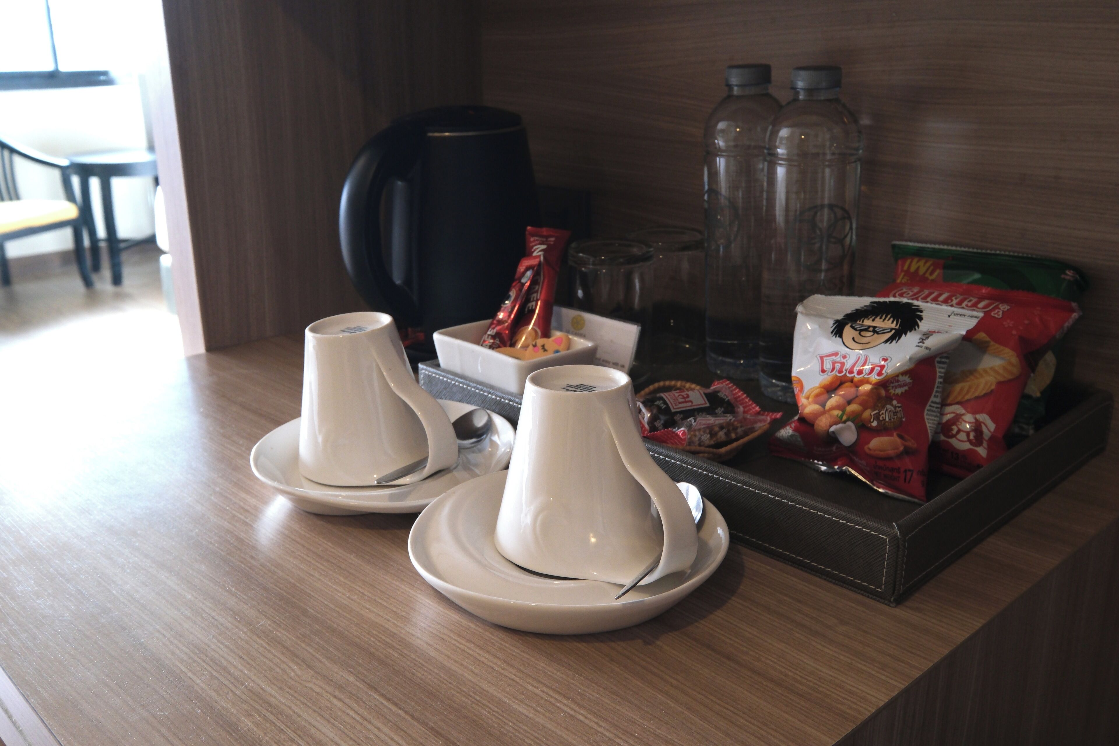 A hotel room's breakfast area with two white teapots on saucers, a tray with snacks, a coffee maker, and water bottles.