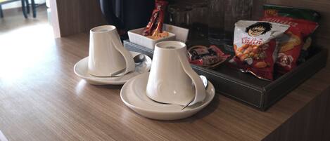 A hotel room's breakfast area with two white teapots on saucers, a tray with snacks, a coffee maker, and water bottles.