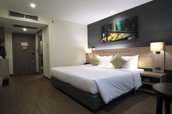 A hotel room with a large bed, two bedside tables, a wall-mounted TV, and a door to the left. - Le Cassia Hotel (Khon Kaen)