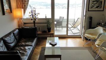 Sala de estar com sofá de couro, mesa de centro de vidro, planta em vaso e vista para uma varanda com mobiliário externo.