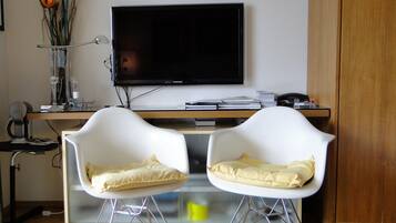Uma sala de estar moderna com uma televisão de tela plana, duas cadeiras brancas, uma escrivaninha com um computador e um vaso com flores.