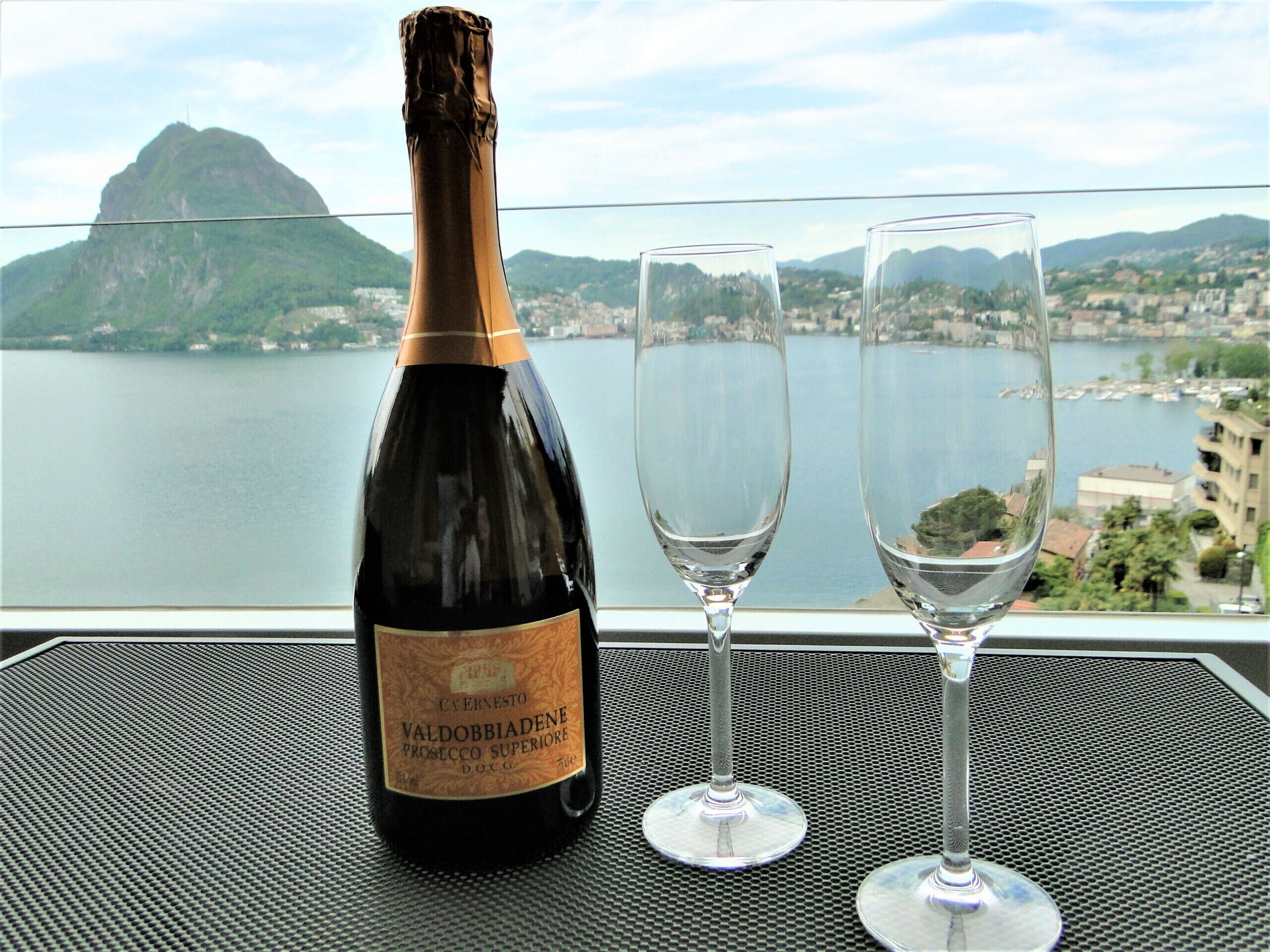A bottle of Valdobbiadene Prosecco Superiore DOCG and two empty wine glasses on a table with a cityscape view.