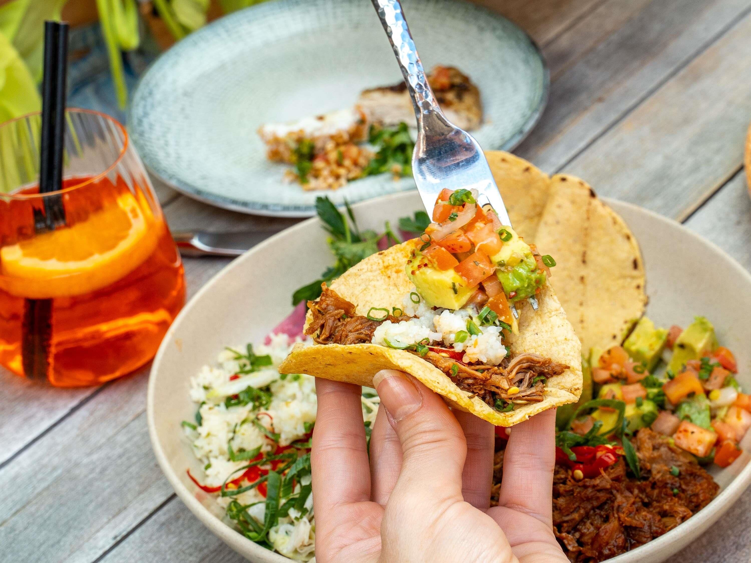 Uma mão segurando um taco com acompanhamentos, uma saladeira e um copo de bebida sobre uma mesa de madeira.