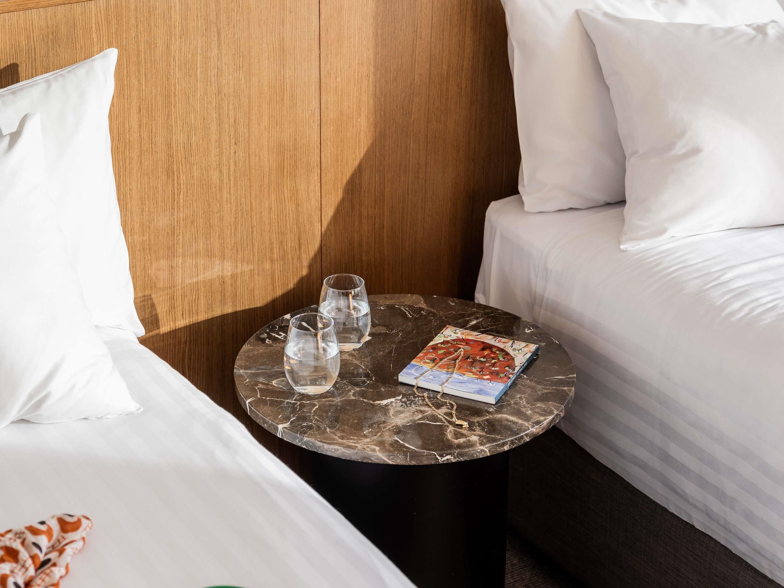 A hotel room with a bed, a small round table with two glasses of water and a magazine, and a wooden headboard.