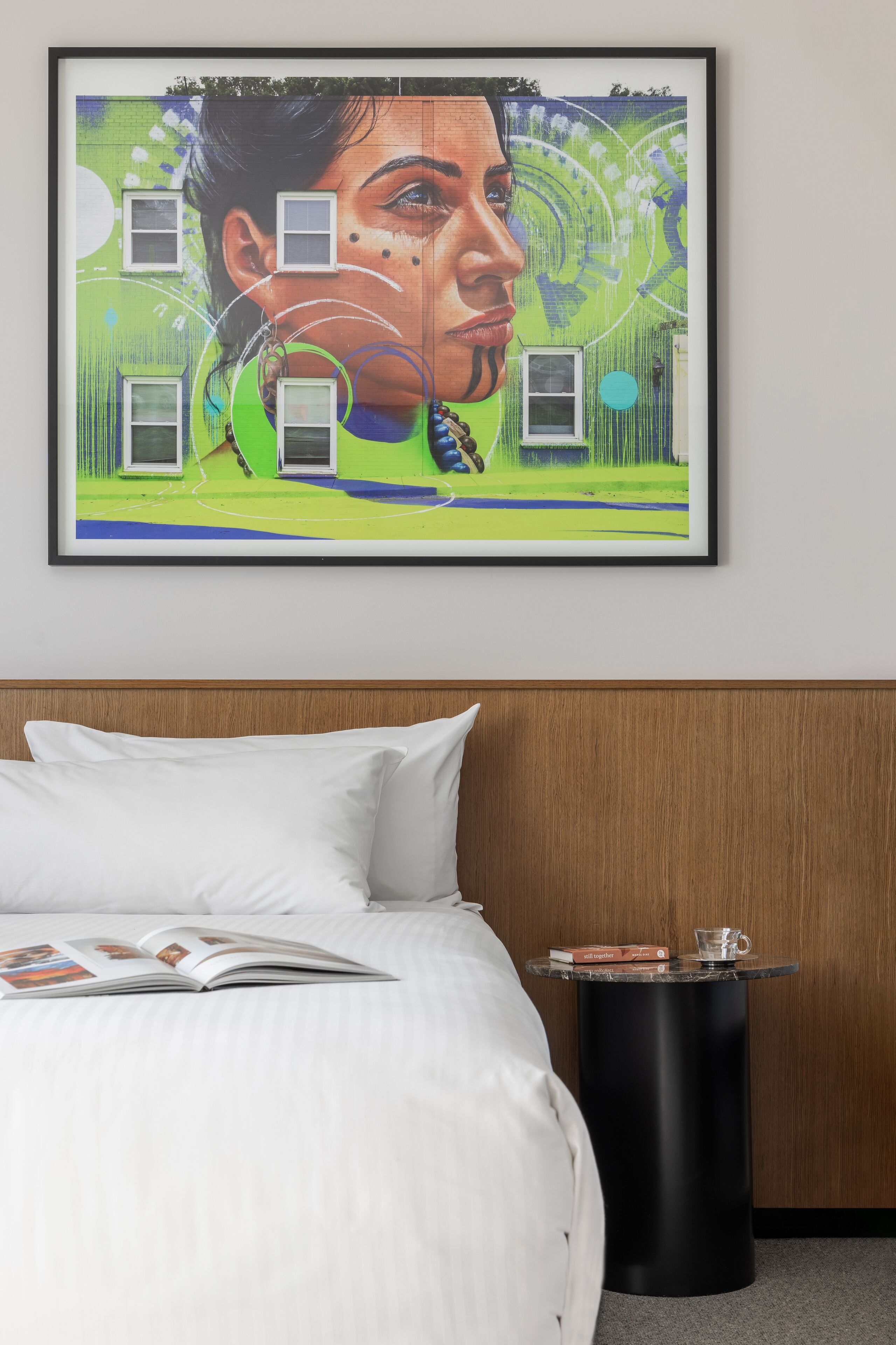 Une chambre d’hôtel avec un lit, une œuvre d’art encadrée au mur, une table de chevet avec un livre et un verre, et une tête de lit en bois.