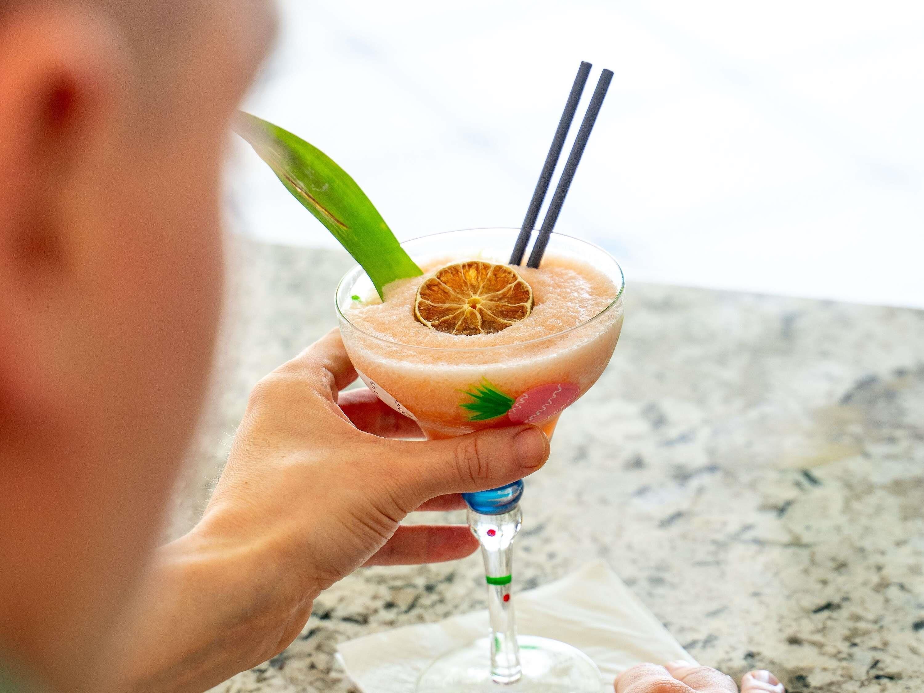 A person holding a cocktail with a straw and a slice of orange garnish.