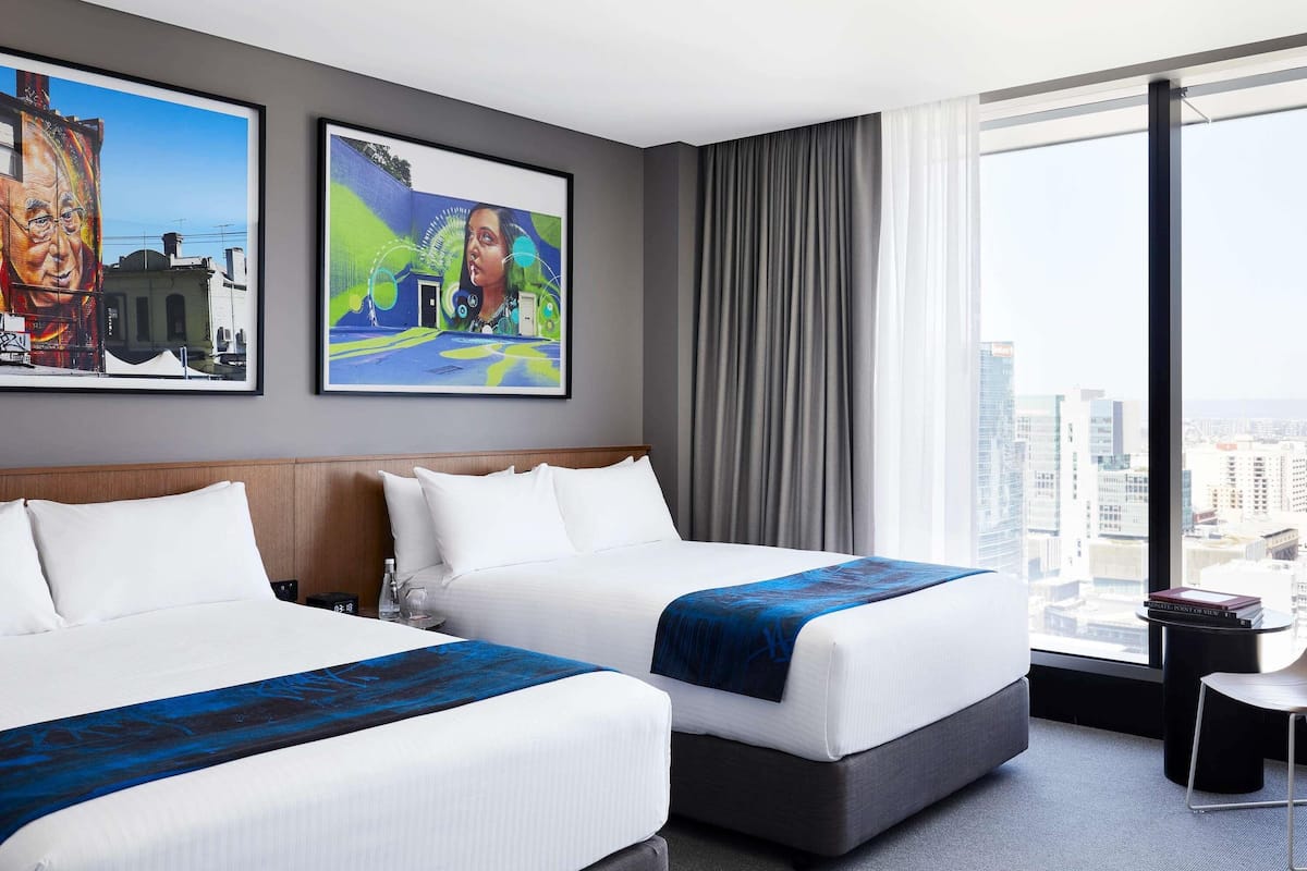 Una habitación de hotel con dos camas, un amplio ventanal con vistas a la ciudad y dos cuadros enmarcados en la pared.