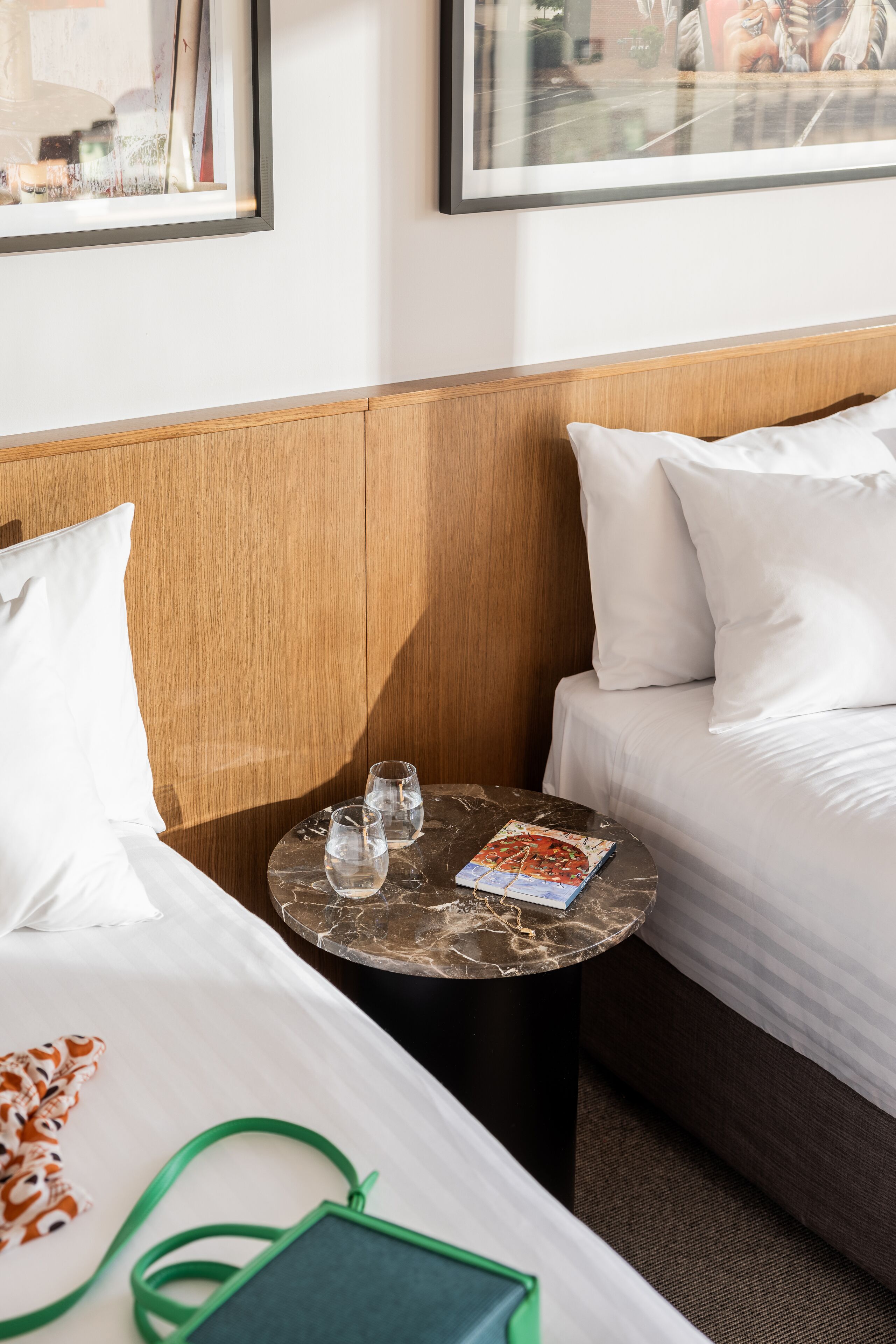 A hotel room with a bed, a small round table with glasses and a magazine, a green bag, and a wooden headboard.