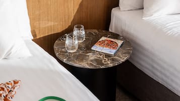 A hotel room with a bed, a small round table with glasses and a magazine, a green bag, and a wooden headboard.