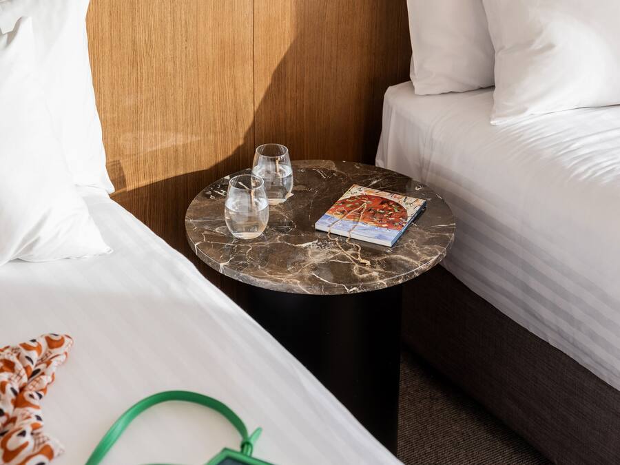 A hotel room with a bed, a small round table with glasses and a magazine, a green bag, and a wooden headboard.