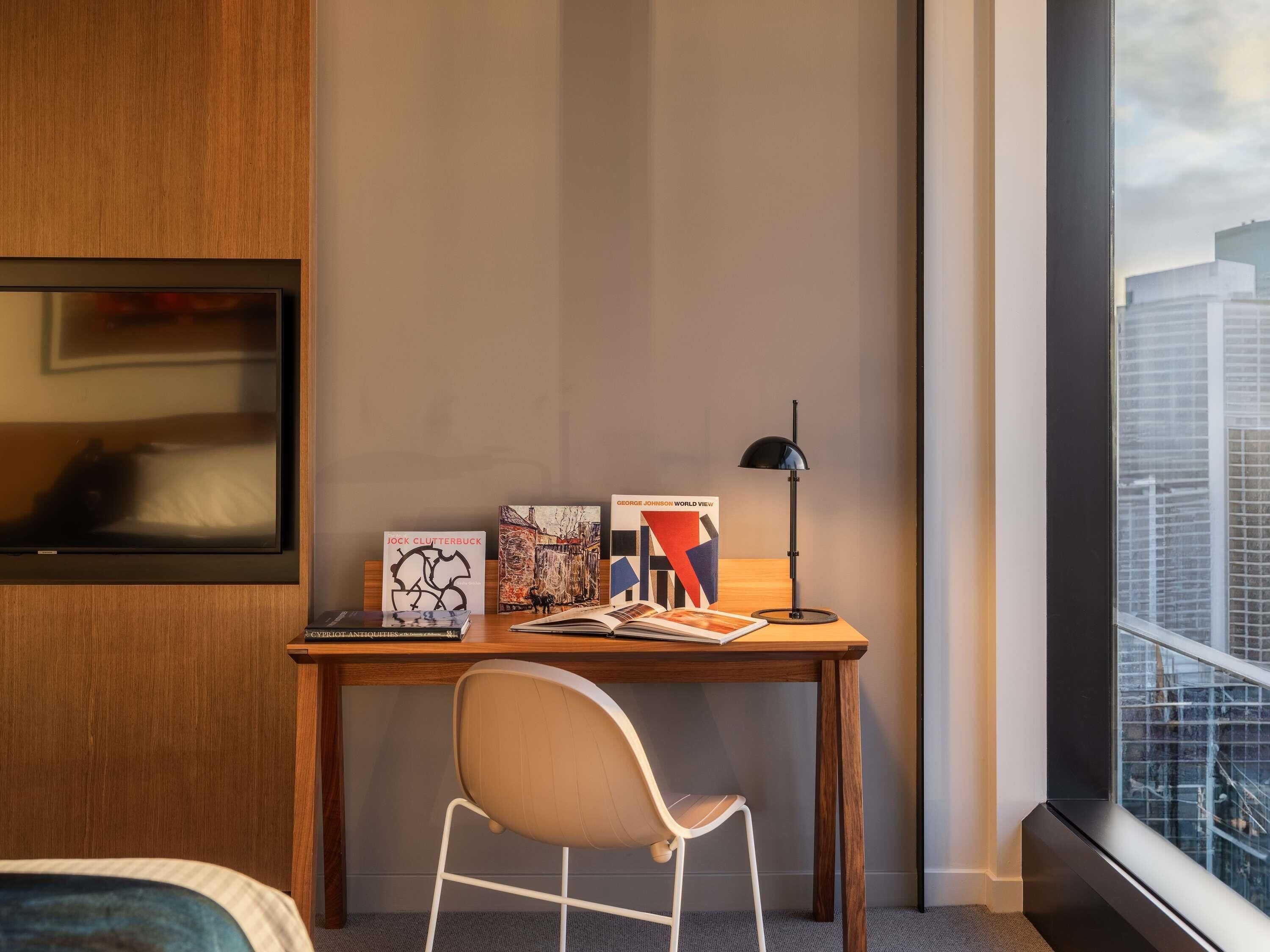 Een moderne hotelkamer met een houten bureau, een witte stoel, een zwarte vloerlamp en kunst aan de muur.